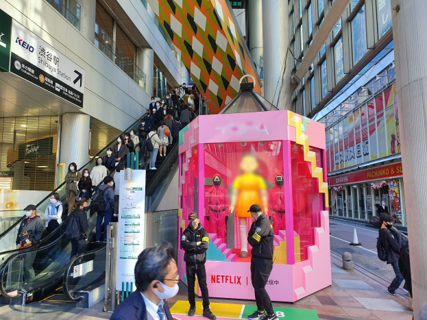 Eine Gruppe von Menschen steht vor einem Gebäude mit Glaswänden, Säulen und Texttafeln, einige tragen Masken, während eine Rolltreppe mit Menschen im Hintergrund zu sehen ist; eine pinkfarbene Box in der Mitte zeigt Schaufensterpuppen und trägt den Text "Netflix's new Netflix booth in Tokyo."