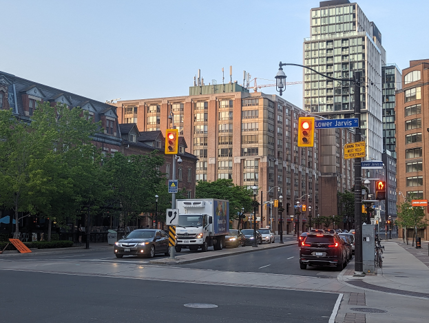 Eine belebte Stadtstraße mit verschiedenen Fahrzeugen, darunter ein Lastwagen, ein Auto und ein SUV, umgeben von Laternenmasten, Ampeln, Texttafeln, Bäumen, Gebäuden mit Fenstern und einem Himmel im Hintergrund.