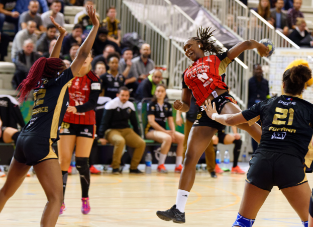 Eine Gruppe von Frauen, die Volleyball auf einem Feld mit einem Ball in der Mitte spielt, während Zuschauer im Hintergrund auf Stühlen, Geländern und Flaschen auf dem Boden sitzen.