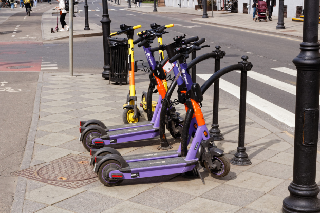 Eine Reihe von Elektrokleinstfahrzeugen, die an einer Straße mit Fußgängern, Radfahrern, einem Müllcontainer im Vordergrund und Gebäuden, Bäumen und Pflanzen im Hintergrund geparkt sind.