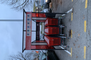 Ein Parkplatz mit mehreren roten Einkaufswagen vor einer Bushaltestelle, Fahrzeuge einschließlich eines Autos und eines Lastwagens auf der Straße und Bäume, Gebäude und einen klaren blauen Himmel im Hintergrund.
