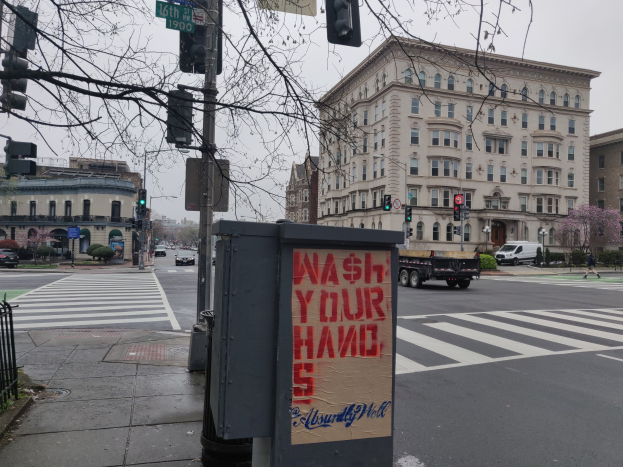 Eine belebte Stadtstraße mit fahrenden Autos, einem Fußgänger auf dem Gehweg, einem Müllcontainer mit der Aufschrift "Hände Waschen", Ampeln, Straßenschilder, einem Metallzaun, Grünpflanzen, Gebäuden und einem klaren blauen Himmel.