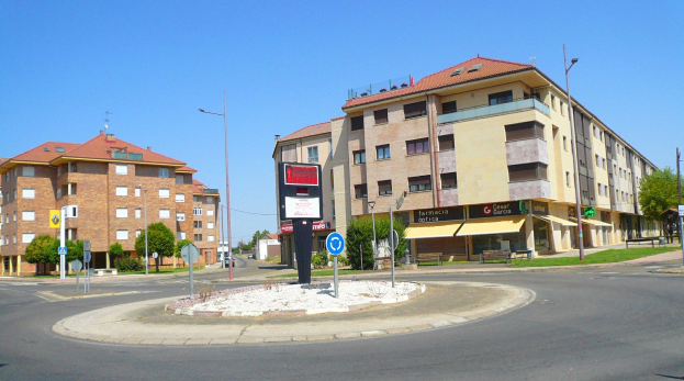 Eine Rundstraße in einer Stadtstraße umgeben von Gebäuden, Straßeninfrastruktur, Fahrzeugen, Grünflächen und einem klaren Himmel.