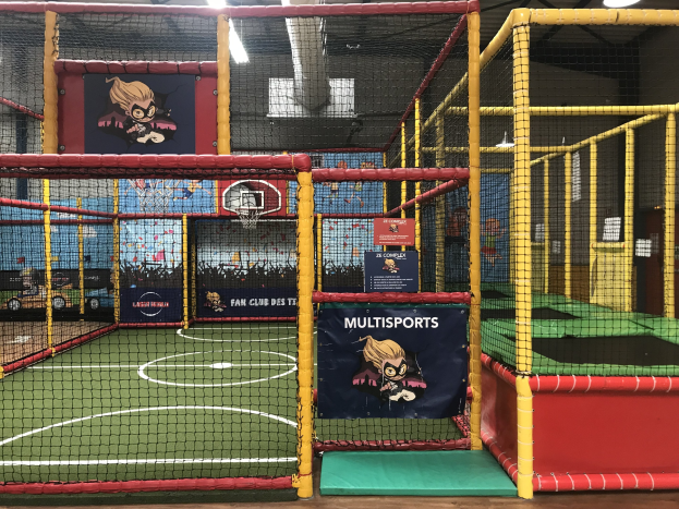 Großes Indoor-Trampolinpark mit einem Basketballfeld in der Mitte, umgeben von einem Netz, mit einem Basketballkorb, Wandbannern und Deckenbeleuchtung.