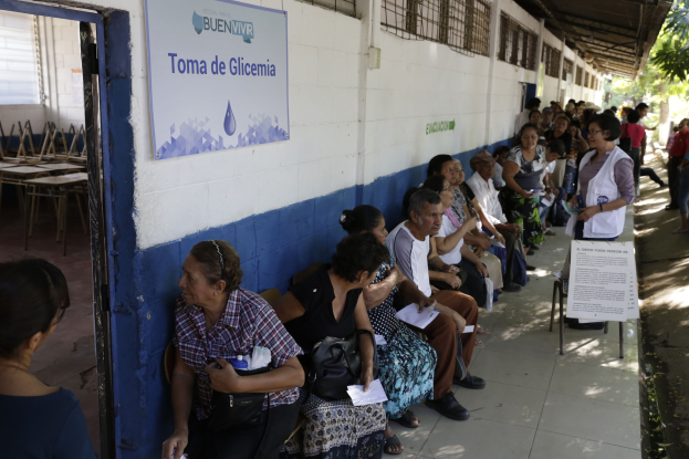 Eine Gruppe von Menschen sitzt auf Bänken vor einem Gebäude, die in einer Schlange auf die Abstimmung an einer Wahllokal in Guadalajara, Mexiko, warten, mit einer Texttafel an der Wand und einer Tür links und Bäumen im Hintergrund.