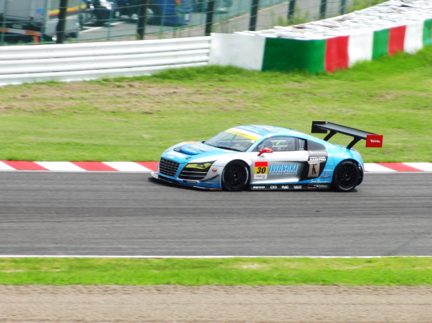 Ein Audi R8 LMS GT3 Rennwagen auf einer Strecke mit Gras auf beiden Seiten, mit einem unscharfen Hintergrund aus einem Zaun, Fahrzeugen und anderen Gegenständen.
