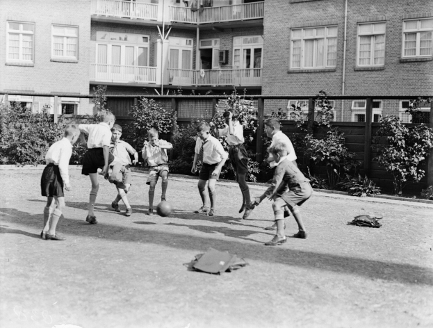 Eine Gruppe von Kindern, die vor einem Gebäude mit Fenstern, Geländern und Balkonen Fußball spielen, mit einem Ball in der Mitte der Gruppe, Pflanzen und einem Zaun im Hintergrund, dargestellt in Schwarz-Weiß.