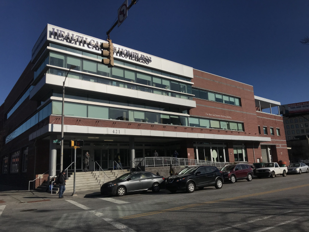 Großes Gebäude, das als New York City Health Care Homeless Center bezeichnet wird, mit geparkten Autos, Fußgängern, Straßenmöbeln und Bäumen unter einem klaren blauen Himmel.