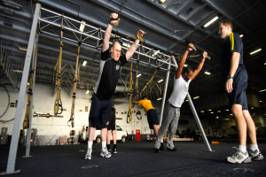 Eine Gruppe von Männern, die Klimmzüge in einem Fitnessstudio machen, wobei ein Mann in der Mitte Fußbekleidung trägt und von Metallstangen, Rahmen, einer Wand, einer Decke mit Deckenlampen und verschiedenen Gegenständen auf der Oberfläche umgeben ist.