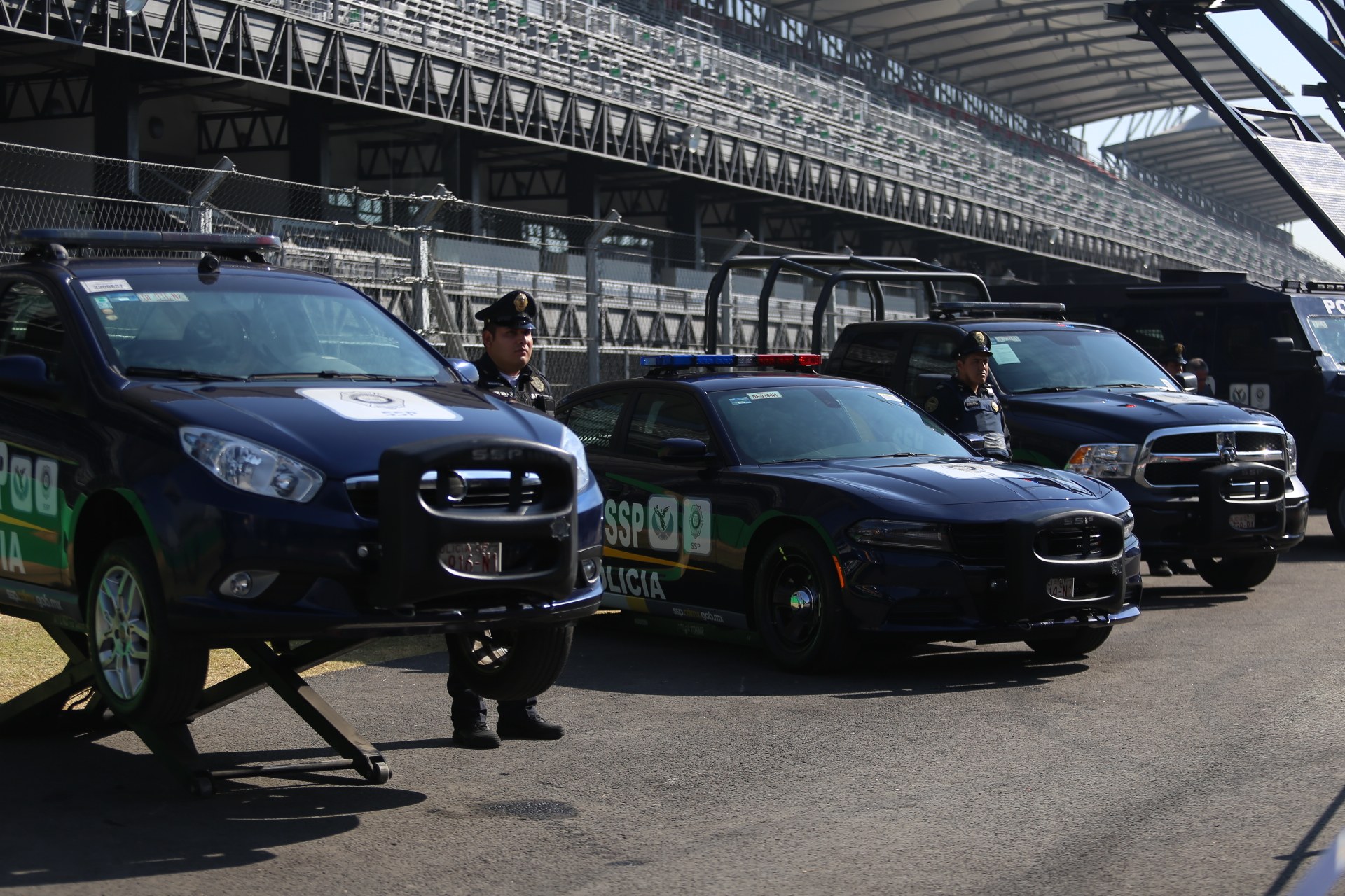 Eine Gruppe von Polizeiwagen vor einer Rennstrecke mit uniformierten Beamten auf der Straße, mit einem Stadion und Maschendrahtzaun im Hintergrund.