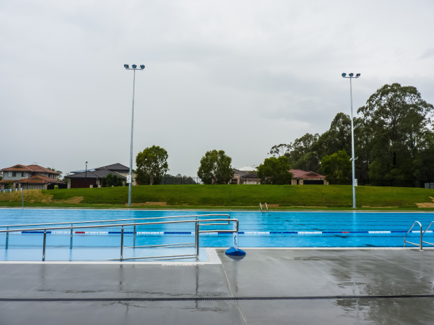 Ein großes Schwimmbad in einem Park, umgeben von Geländern, Pfählen, Laternen und Bäumen, mit Häusern und einem klaren blauen Himmel im Hintergrund.