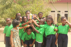 Gruppe von Kindern mit Pokal in einer Schulumgebung mit Bäumen und einem Gebäude im Hintergrund.