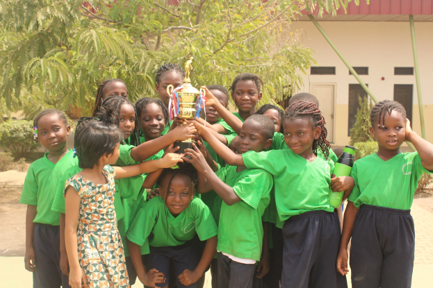 Gruppe von Kindern mit Pokal in einer Schulumgebung mit Bäumen und einem Gebäude im Hintergrund.