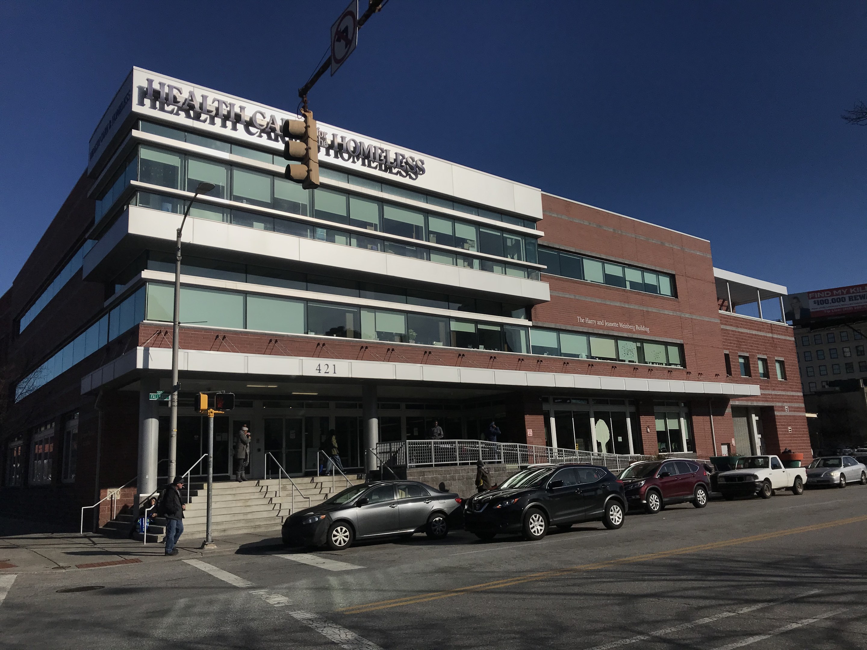 Großes Gebäude mit der Aufschrift "New York City Health Care Homeless Center" mit parkenden Autos, Passanten, Straßenmöblierung und Bäumen unter einem klaren blauen Himmel.
