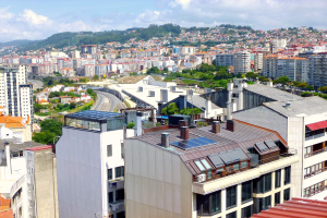 Eine Stadtansicht von einem hohen Standpunkt aus, die viele Gebäude, Bäume, Straßen, Fahrzeuge und entfernte Berge unter einem bewölkten Himmel zeigt, wobei auf dem Dach eines Gebäudes Solarpanels zu sehen sind.