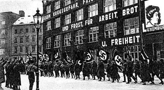 Schwarzes und wei├čes Bild einer Gruppe, die mit Fahnen und Transparenten die Stra├če entlangmarschiert, mit einem Lichtmast links und einer Tafel mit der Aufschrift "Deutsche Nationalsozialistische Arbeiterpartei in Berlin" rechts.