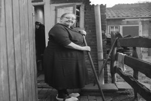 Eine ältere Frau in einem Kleid und Brille steht vor einem Haus und hält einen Stock in der Hand, rechts neben ihr ist ein Holzzaun und im Hintergrund sind andere Häuser zu sehen.