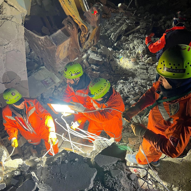 Eine Gruppe von Menschen in orangen Uniformen und grünen Helmen bei der Arbeit an Schutt von einem eingestürzten Gebäude mit einem Kran im Hintergrund.