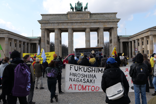 Demonstranten halten Schilder und Fahnen mit der Aufschrift 'Fukushima Hiroshima Nagasaki Atomod' vor dem Brandenburger Tor in Berlin, Deutschland, wobei die Säulen und Statuen des Monuments im Hintergrund zu sehen sind.