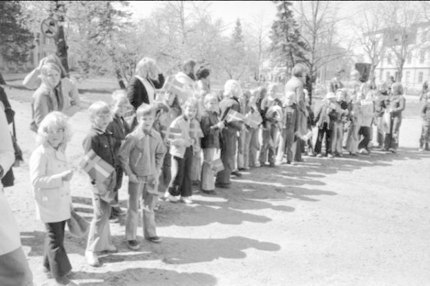 Eine Gruppe von Menschen steht in einer Reihe auf einem Schotterweg und hält Fahnen, mit Bäumen, Gebäuden und einem klaren Himmel im Hintergrund, auf einem Schwarz-Weiß-Bild einer Protestkundgebung auf dem Schulgelände.