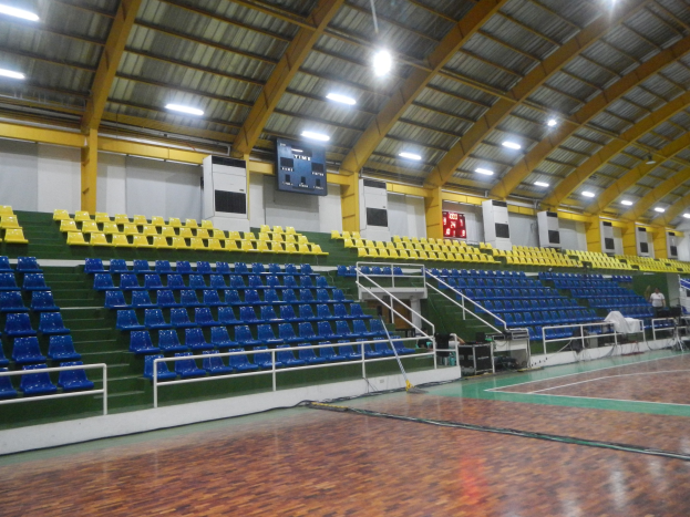 Großer Hallensportplatz mit Holzboden, blauen und gelben Sitzplätzen, Geländern, Treppen zum Sitzbereich, Wandbildschirme, Deckenleuchten und einigen Personen.