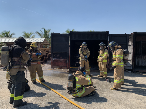 Eine Gruppe von Feuerwehrleuten in Helmen und Zylindern steht vor einem Gebäude, hält eine Leitung und hat Bäume, Schuppen und hölzerne Gegenstände im Hintergrund, während einer Feuerlöschübung.