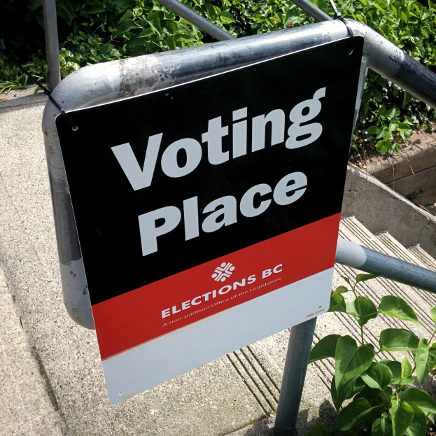 Ein Wahlplatzschild an einem Gebäude mit einer Tafel an einem Geländer im Vordergrund und Pflanzen im Hintergrund.