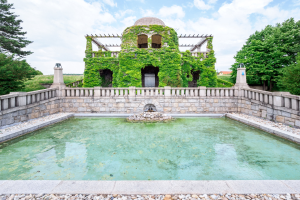 Das Hiroshima-Friedensdenkmal, ein großes Gebäude mit Efeu an seiner Seite, umgeben von einer Wand und einem zentralen Brunnen, mit Steinen im Vordergrund und Bäumen und einem bewölkten Himmel im Hintergrund.