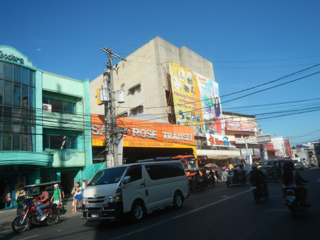 Eine belebte Stadtstraße mit zahlreichen Autos, Motorrädern, Fußgängern, Radfahrern und einem geparkten Kleinbus, sowie Gebäuden, Werbetafeln, Strommasten mit Drähten und einem klaren blauen Himmel.