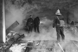 Eine Gruppe von Feuerwehrmännern in Helmen und mit Taschen auf den Schienen stehend, mit Rauch, der von links aufsteigt und verstreuten Gegenständen auf dem Boden, dargestellt in Schwarz-Weiß.