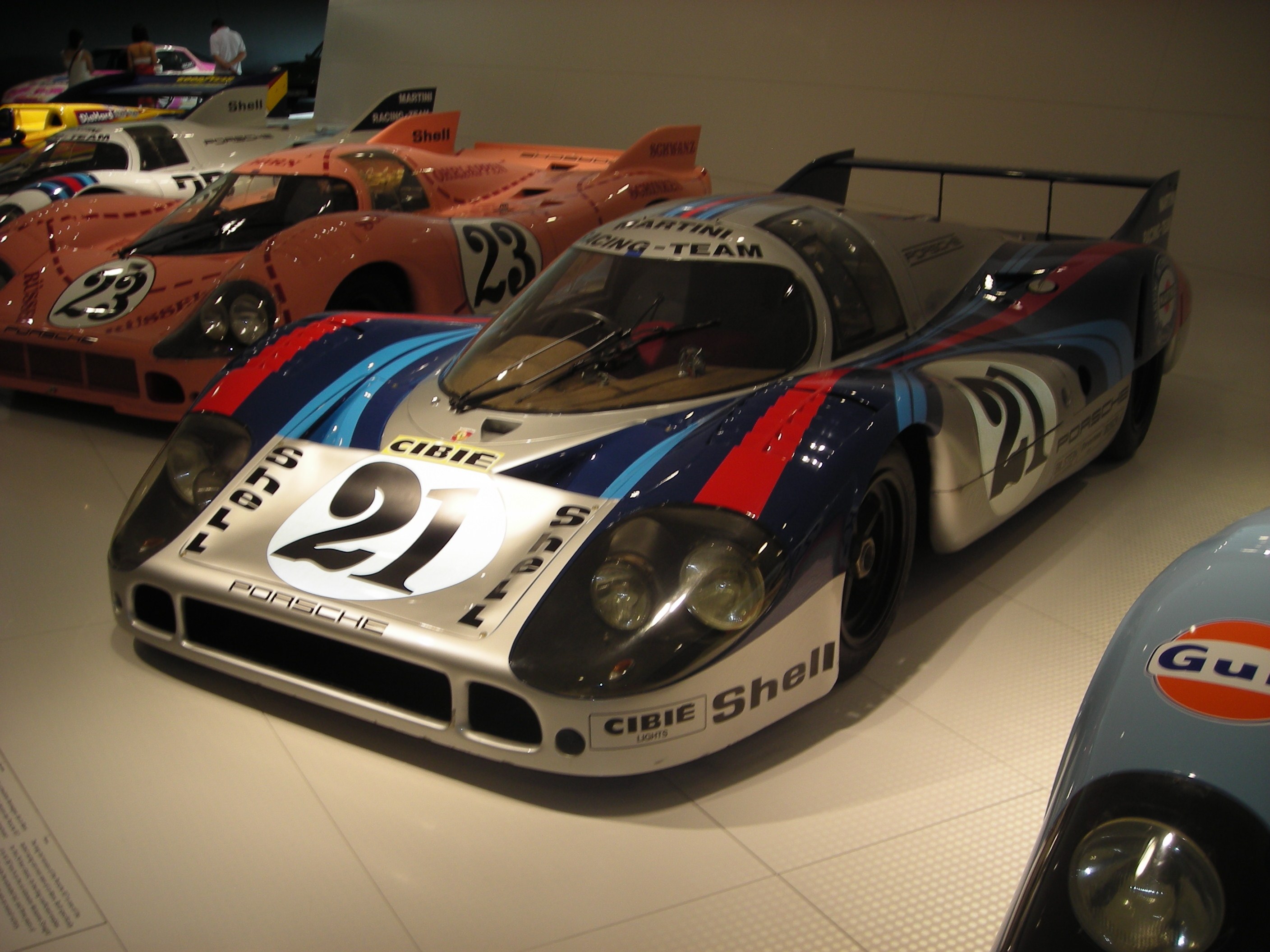 Ein Porsche 917K Le Mans Rennauto in einem Museum ausgestellt, umgeben von anderen Sportwagen, mit Menschen in der Nähe und einer Wand im Hintergrund; ein Blatt mit Text ist auf der linken Seite sichtbar.