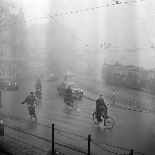 Schwarzes und weißes Foto von Menschen, die auf der Straße Fahrräder fahren, mit Fahrzeugen, Kettenpfählen und Gebäuden im Hintergrund.