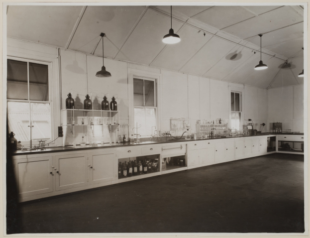 Schwarz-weiß-Foto eines Labors mit einer langen Theke, Schäben, Regalen mit Flaschen, Fenstern, Deckenlampen und verschiedenen Gegenständen auf der Theke.