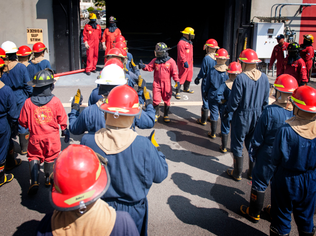 Eine Gruppe von Feuerwehrleuten in roten Uniformen und Helmen, die eine Straße entlanggehen, mit einer Wand auf der linken Seite und Bäumen und einem klaren blauen Himmel im Hintergrund.