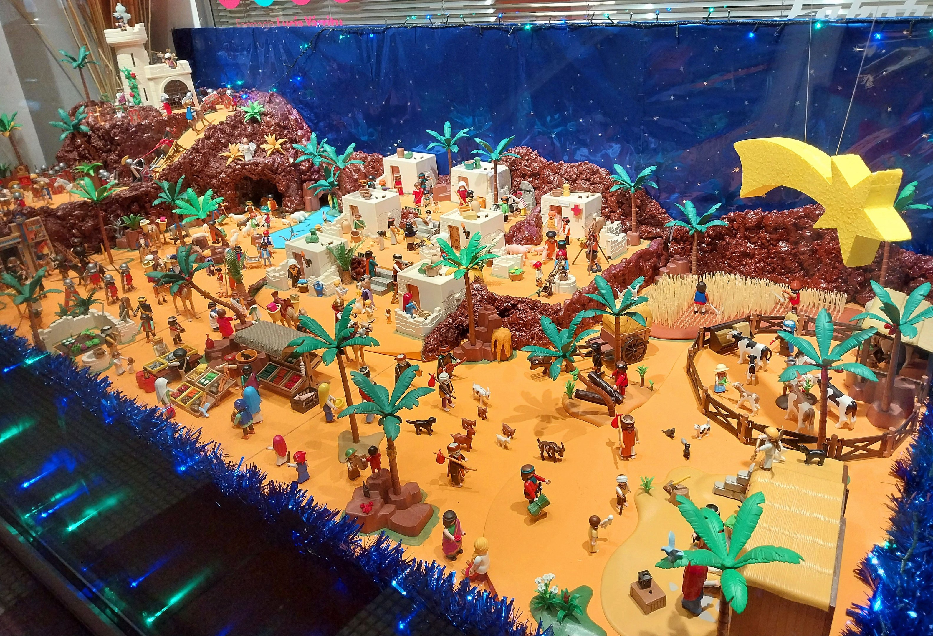 Vitrine mit verschiedenen Lego-Steinen und -Teilen auf einem Tisch, umgeben von Spielzeugen, Bäumen und dekorativen Gegenständen, mit einer beleuchteten Wand im Hintergrund.