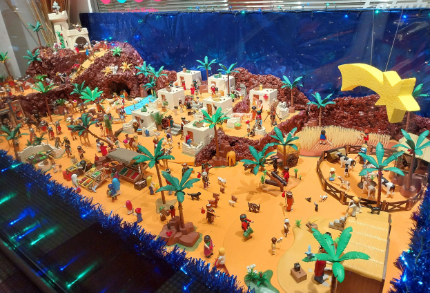 Vitrine mit verschiedenen Lego-Steinen und -Teilen auf einem Tisch, umgeben von Spielzeugen, Bäumen und dekorativen Gegenständen, mit einer beleuchteten Wand im Hintergrund.