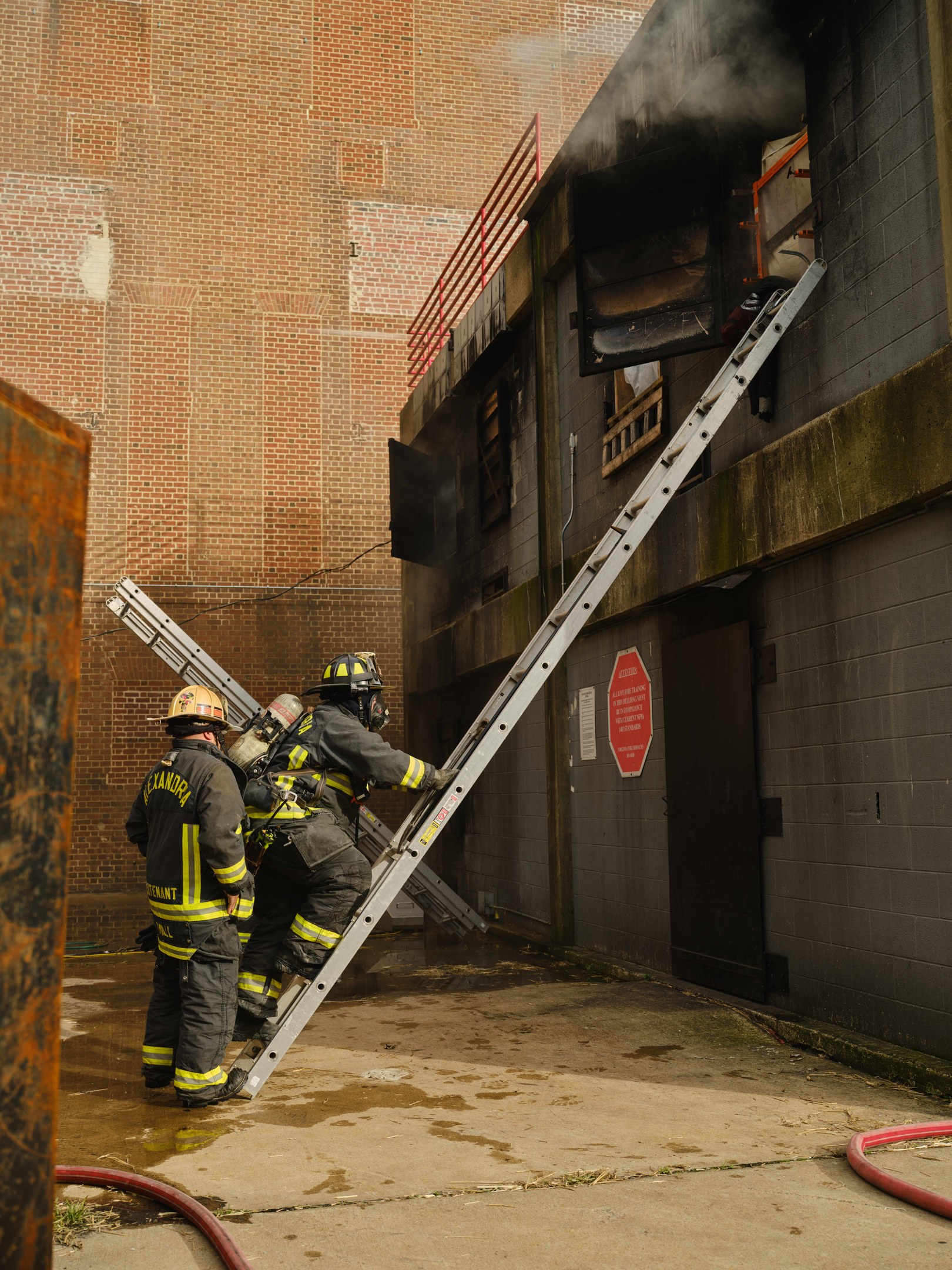 Feuerwehrleute in Helmen klettern an einer Leiter zu einem raucherfüllten Gebäude, mit einem Metallobjekt links und Rohren am Boden.