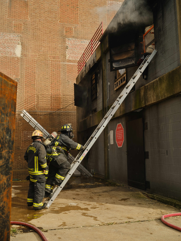 Feuerwehrleute in Helmen klettern an einer Leiter zu einem raucherfüllten Gebäude, mit einem Metallobjekt links und Rohren am Boden.