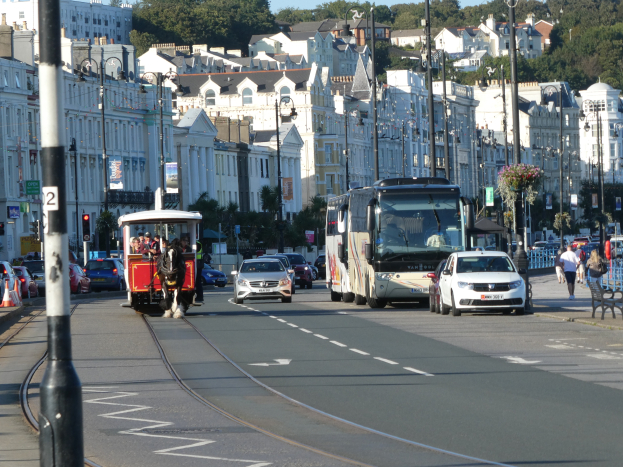 Eine belebte Stadtstraße mit zahlreichen Fahrzeugen, Fußgängern, hohen Gebäuden, Verkehrsampeln, Laternen, Texttafeln und Bäumen unter einem klaren blauen Himmel.