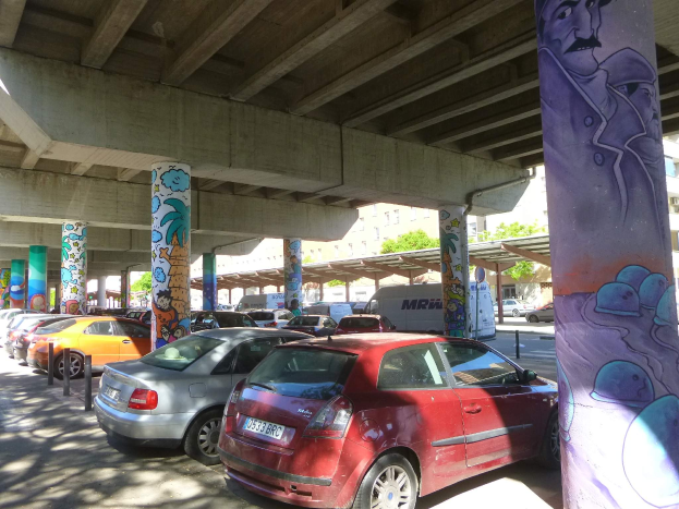 Ein Parkplatz voller parkender Autos unter einer Brücke, mit Graffiti an den Pfeilern und Gebäuden, mit Bäumen im Hintergrund und einem Van auf dem Parkplatz.