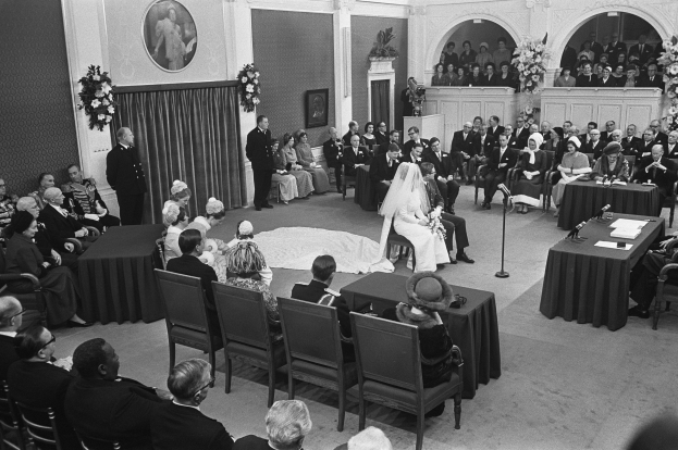 Ein Schwarz-Weiß-Foto einer Hochzeitszeremonie in einem großen Raum mit Gästen auf Stühlen und Tischen mit Papieren und Mikrofonen, einem Podium mit Mikrofonständer und einer Wand mit Foto-Rahmen, Blumenbouquets und einem Vorhang.