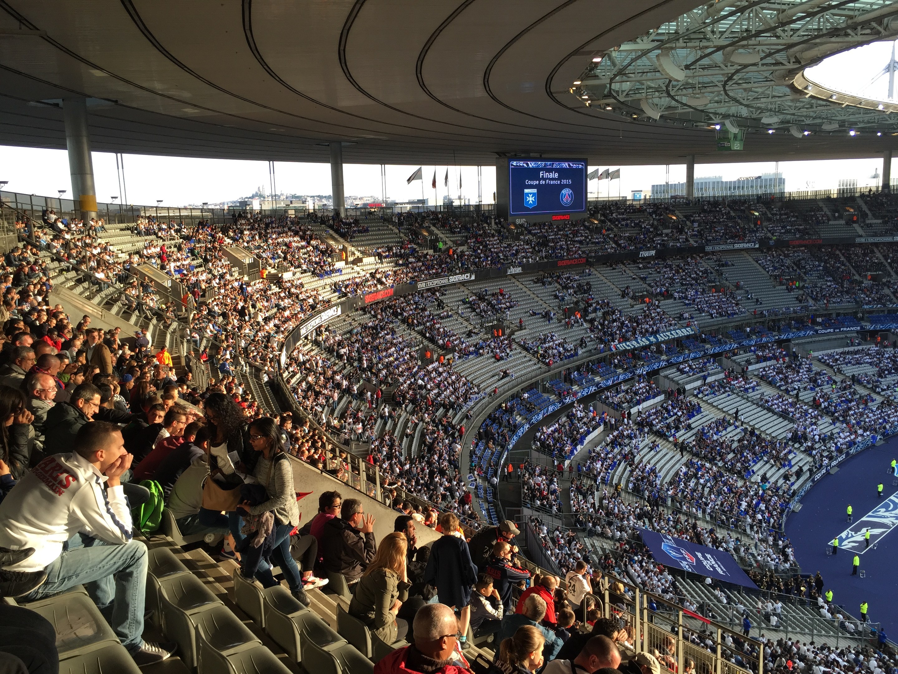 Große Menschenmenge in einem Stadion bei einem Füßtballspiel mit einer Bühne, Fahnen, Masten und einem Bildschirm im Hintergrund, identifiziert als Allianz Arena in München, Deutschland.