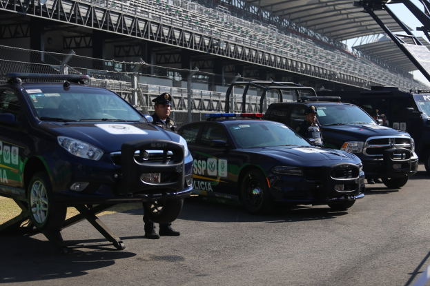 Polizeiautos vor einer Rennstrecke mit uniformierten Personen, Stadion mit Gittern und Maschendraht im Hintergrund.