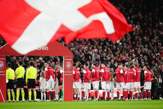 Gruppe von Menschen auf einem Fußballfeld mit einer roten und weißen Fahne im Vordergrund, einem Bogen mit der Aufschrift "Bayern München vs. Bayern München Wett-Tipps & Vorschau" im Hintergrund und einer großen Menge im Stadion.