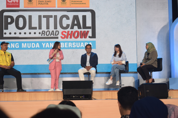 Eine Gruppe von Menschen sitzt auf einer Bühne mit einer Frau in der Mitte, die ein Mikrofon hält, einem Publikum im Vordergrund und einem Banner im Hintergrund mit der Aufschrift 'Politische Straßen-show 2019'