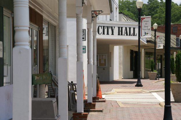 Rathaus mit Gebäuden, Straßenlaternen, Straßenschildern, Kraftfahrzeugen, Absperrgittern, Stühlen, Topfpflanzen, Bäumen und einer bewölkten Himmel im Hintergrund.