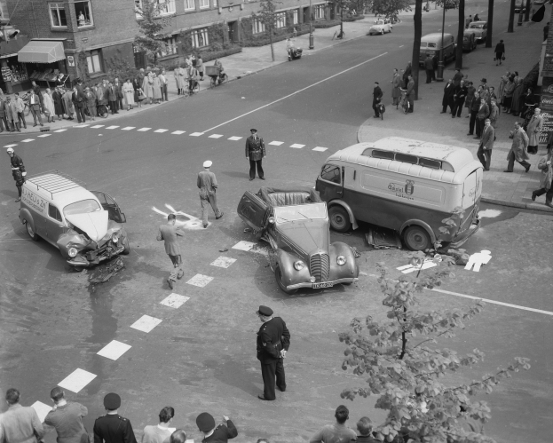 Schwarz-weißes Bild eines Autounfalls an einer Straßenkreuzung mit mehreren Fahrzeugen, darunter ein Van, und einer Gruppe von Schaulustigen; Gebäude, Bäume, Pfosten und Texttafeln sind im Hintergrund zu sehen.