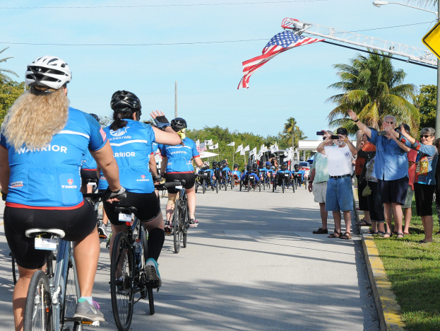 Radfahrer mit Helmen fahren eine von Bäumen gesäumte Straße während eines Triathlon-Events, mit Zuschauern auf dem Rasen, die Fotos und Videos aufnehmen, vor einem klaren blauen Himmel.