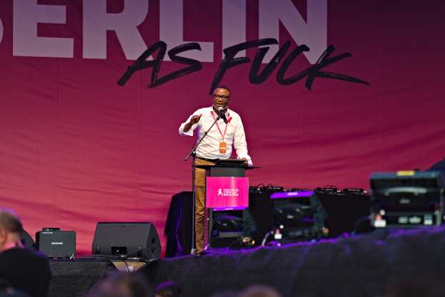 Ein Mann in weißem Hemd und braunen Hosen steht auf einer Bühne und hält ein Mikrofon in der Hand, hinter ihm eine pinkfarbene Fahne mit der Aufschrift 'Berlin as Fuck' und Lautsprecher auf der Bühne.