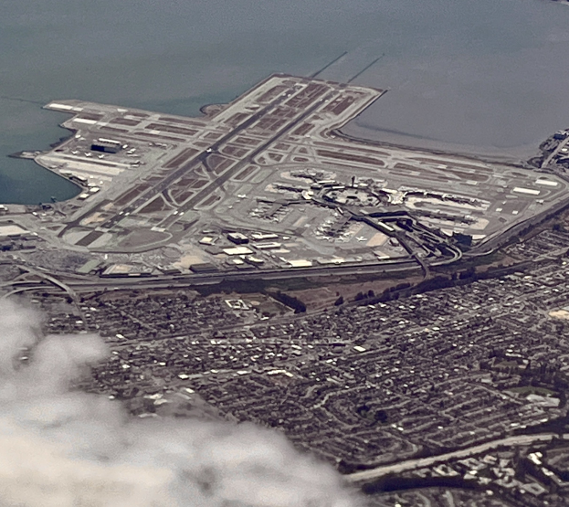 Luftaufnahme eines Flughafens mit Gebäuden, Straßen, Bäumen und Wasser sowie einem Hauch von Rauch am Boden.